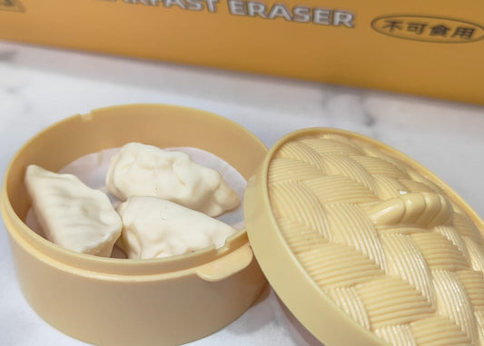 A beige plastic steamer dumpling mold set, showing the open round mold filled with three dumplings, its textured lid leaning against it, and a yellow box in the blurred background on a white marble surface.